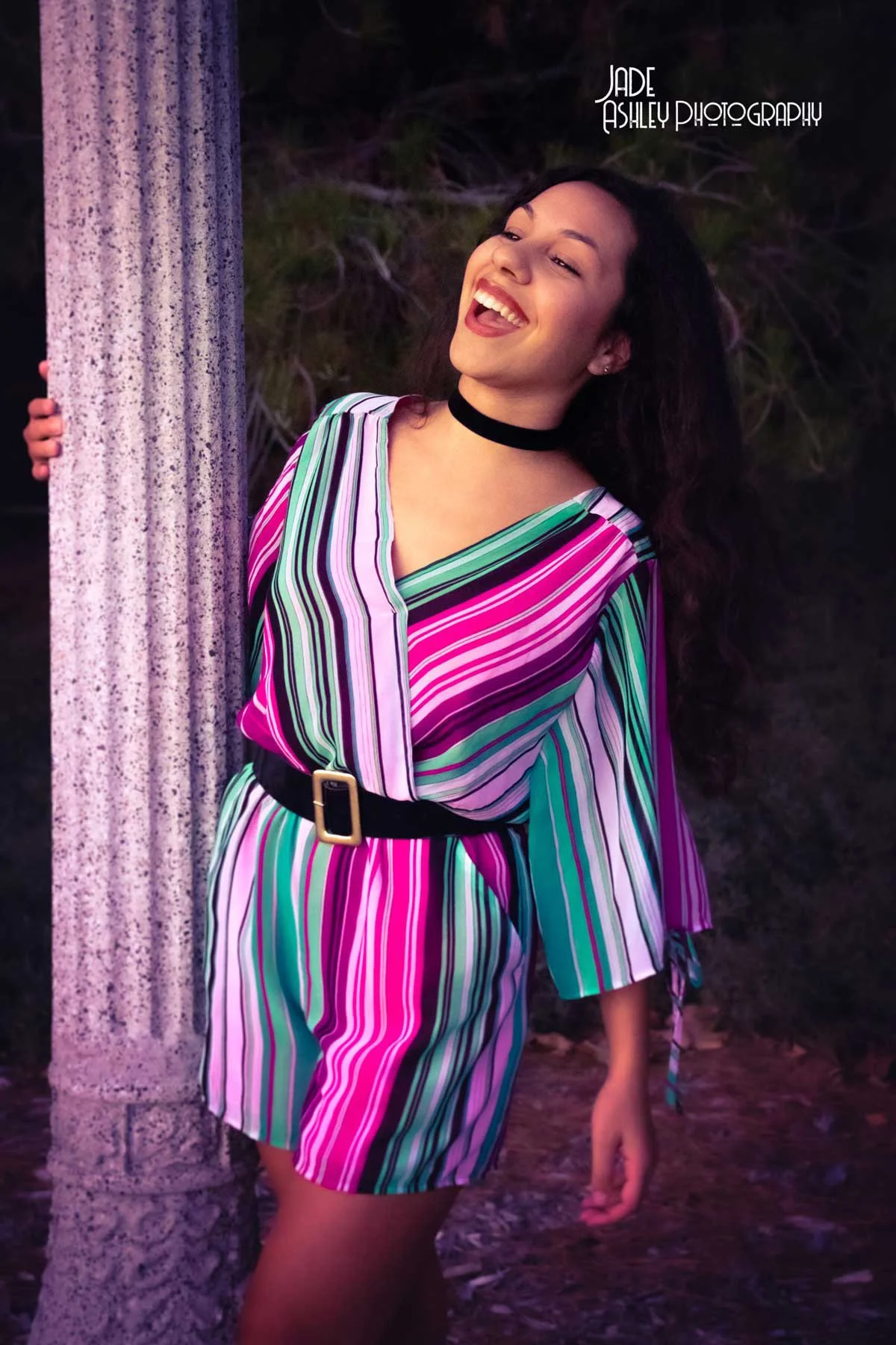 Young woman smiling and laughing while standing outdoors next to a textured concrete pillar, wearing a colorful, striped dress and a black choker necklace.