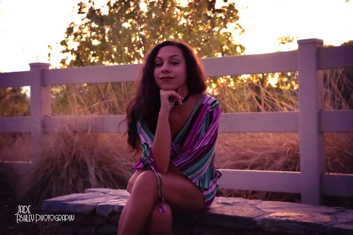 A woman sitting outdoors on a stone ledge during sunset, wearing a colorful striped dress and a choker necklace, with her chin resting on her hand.