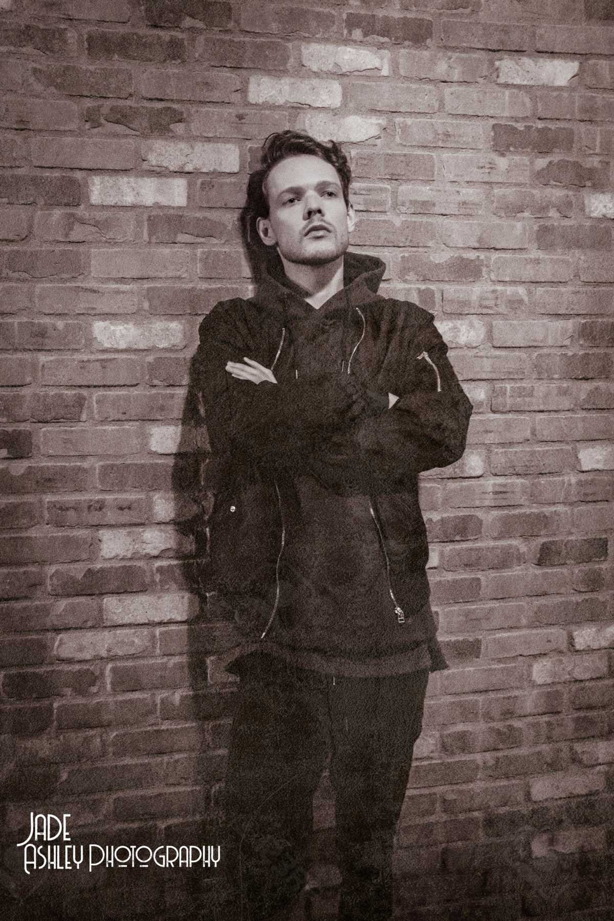 Black and white photo of a young man with styled hair and facial hair, standing against a brick wall with his arms crossed, wearing a dark jacket and hoodie, looking confidently at the camera.