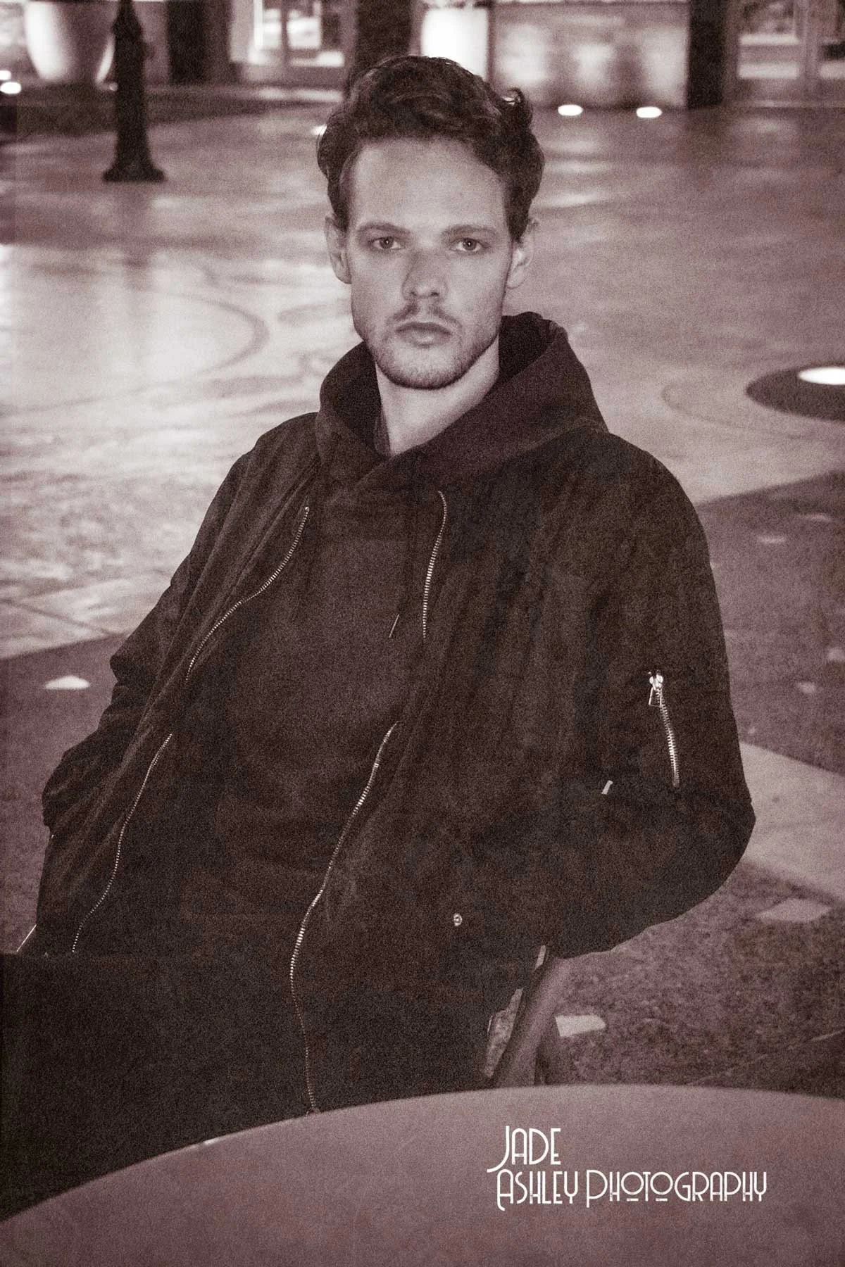 A young man with curly hair and a beard sitting outdoors at night, wearing a dark hoodie and jacket. The setting appears to be an urban area with a sidewalk and streetlights in the background. The photo is in black and white. The bottom right corner 