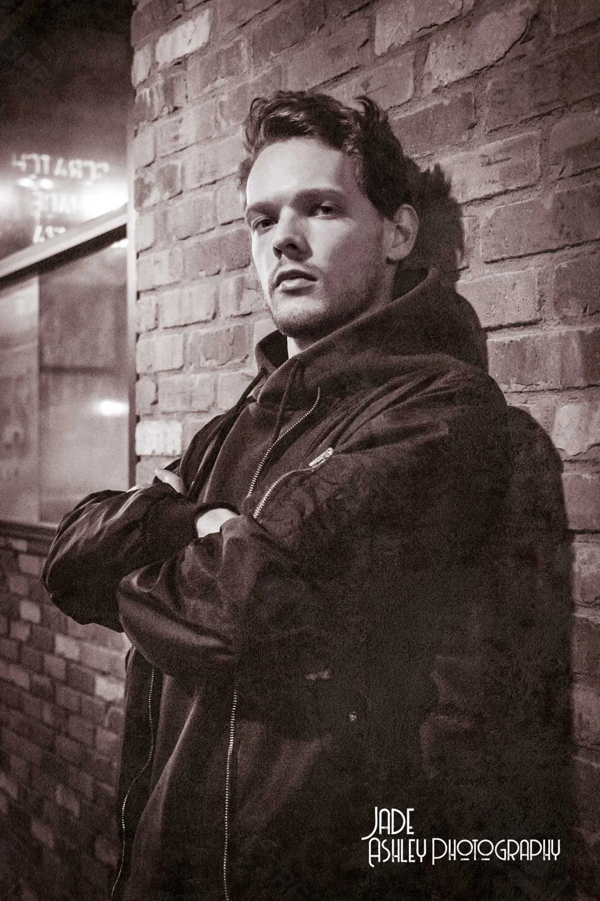 A young man with short dark hair and light skin leaning against a brick wall in a black leather jacket with crossed arms, gazing at the camera in a dimly lit setting.
