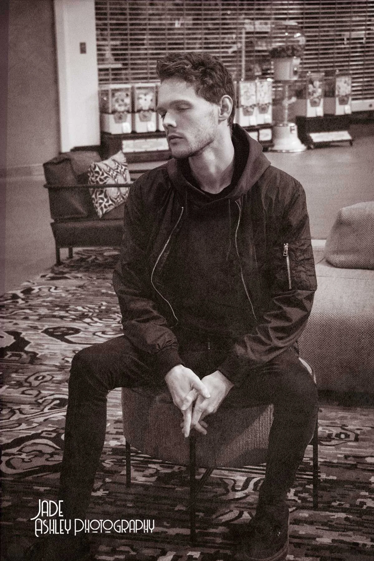 A young man with dark hair, wearing a black jacket and black pants, sitting on a stool in a room with a patterned rug, a sofa, and a background with shelves and storage boxes.