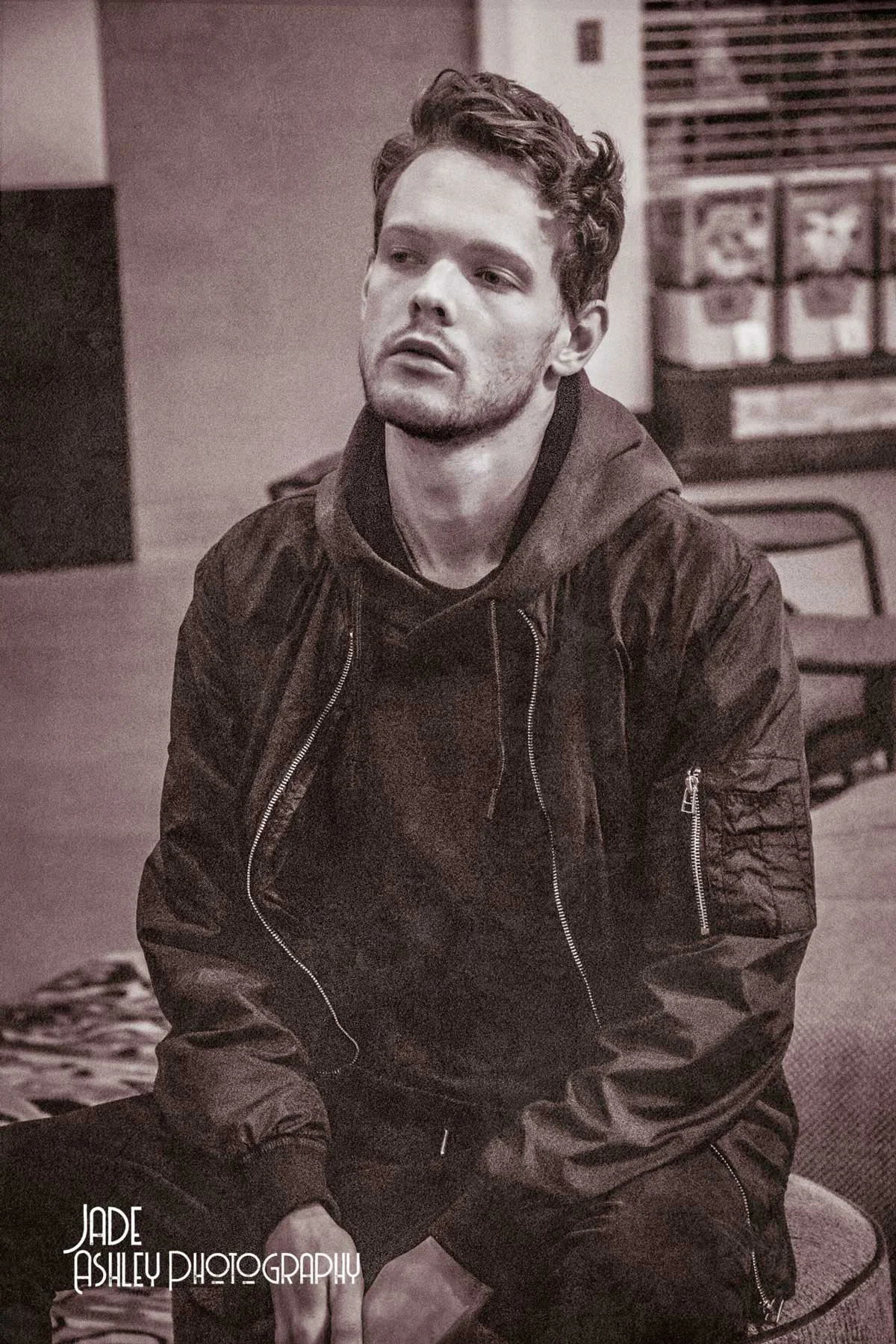 A young man with curly hair and a beard, wearing a dark jacket and hoodie, sitting indoors with a serious expression, in front of shelves with storage bins.