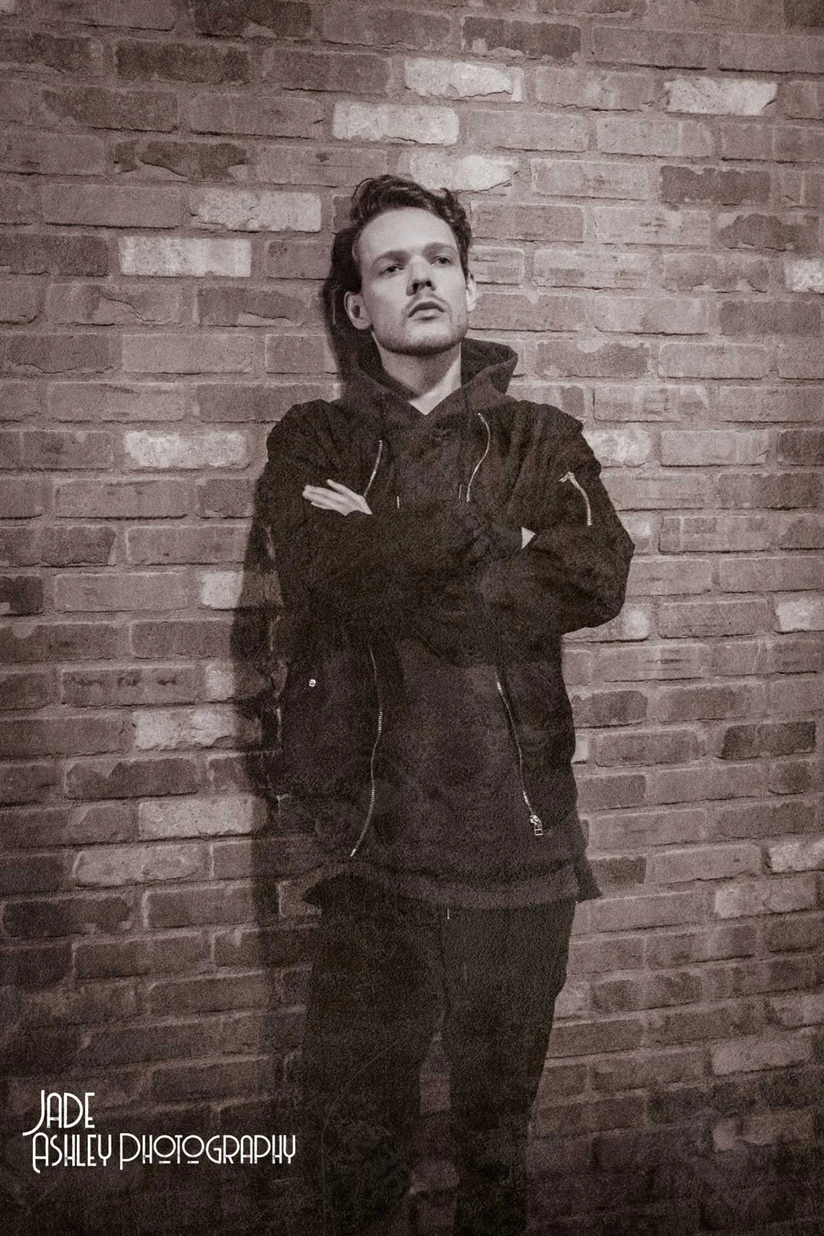 A young man with styled hair and a serious expression, standing with arms crossed against a brick wall, wearing a dark hoodie and jacket.