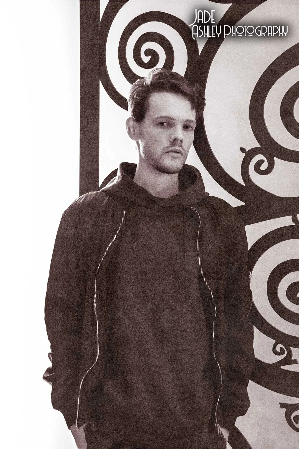 A young man with dark hair and facial hair, wearing a dark hoodie and jacket, stands in front of a black and white patterned background with spiral designs. The text "Jade Ashley Photography" appears in the upper right corner.