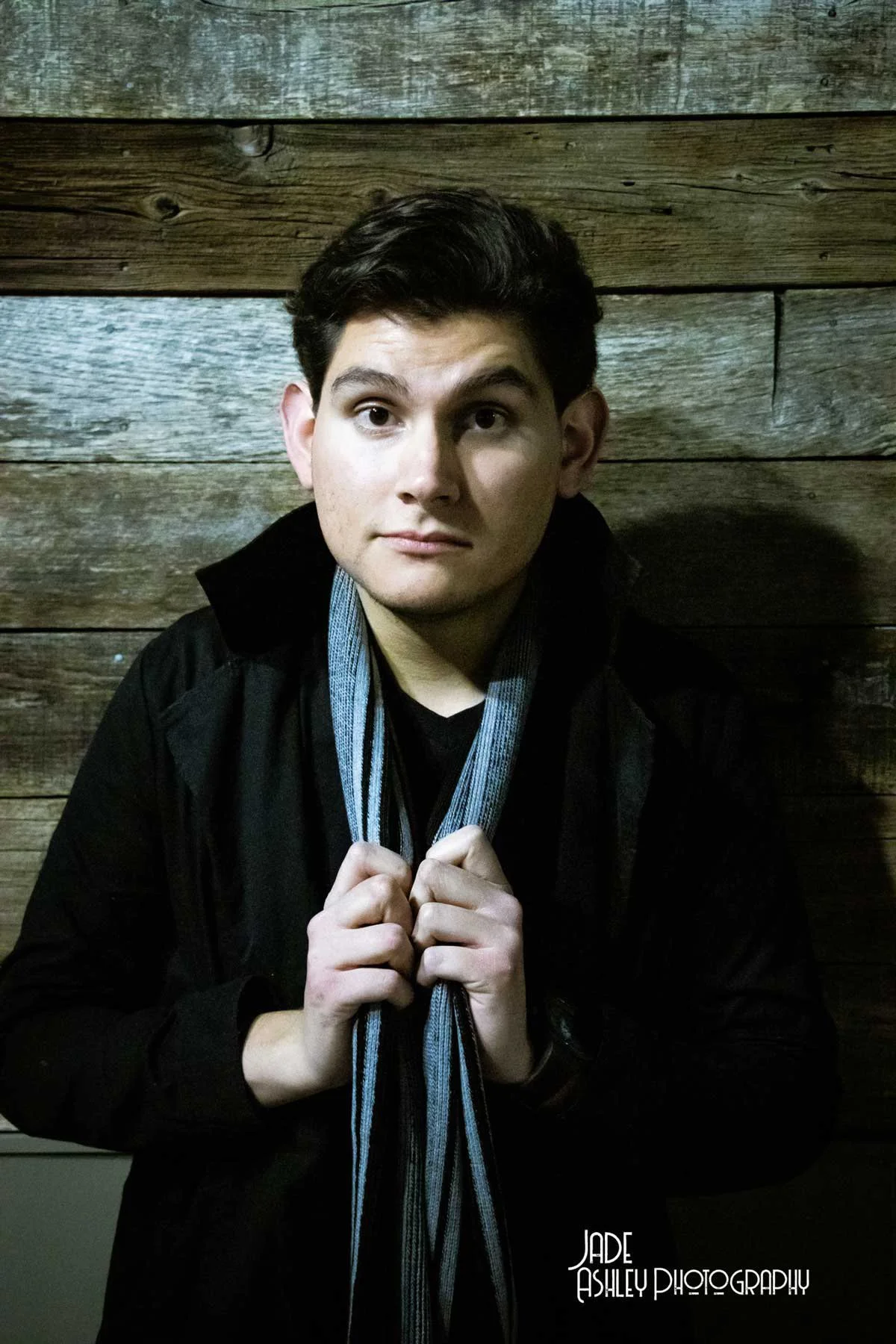 Young man with dark hair wearing a black jacket and striped scarf, standing against a wooden wall.
