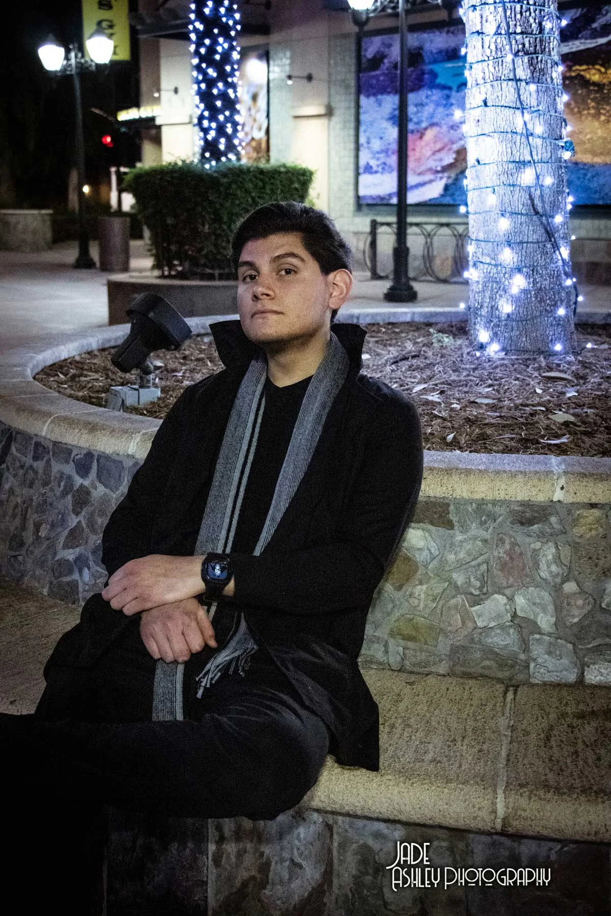 A young man sitting on a stone bench in a well-lit outdoor area at night. He has dark hair, is dressed in black, and wears a gray scarf and a watch. Behind him, there are trees wrapped in blue lights, a bush, and a building with street lamps and a di