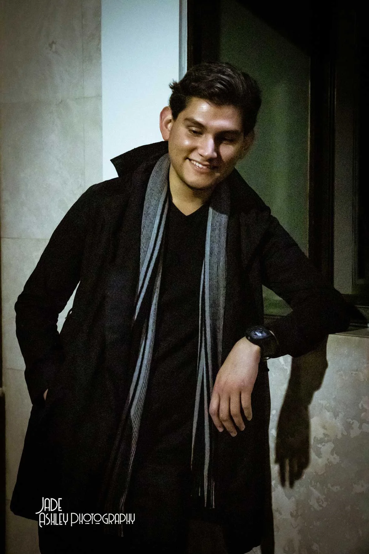 A smiling young man with dark hair, wearing a black coat, a black shirt, and a gray striped scarf, leaning against a wall indoors.