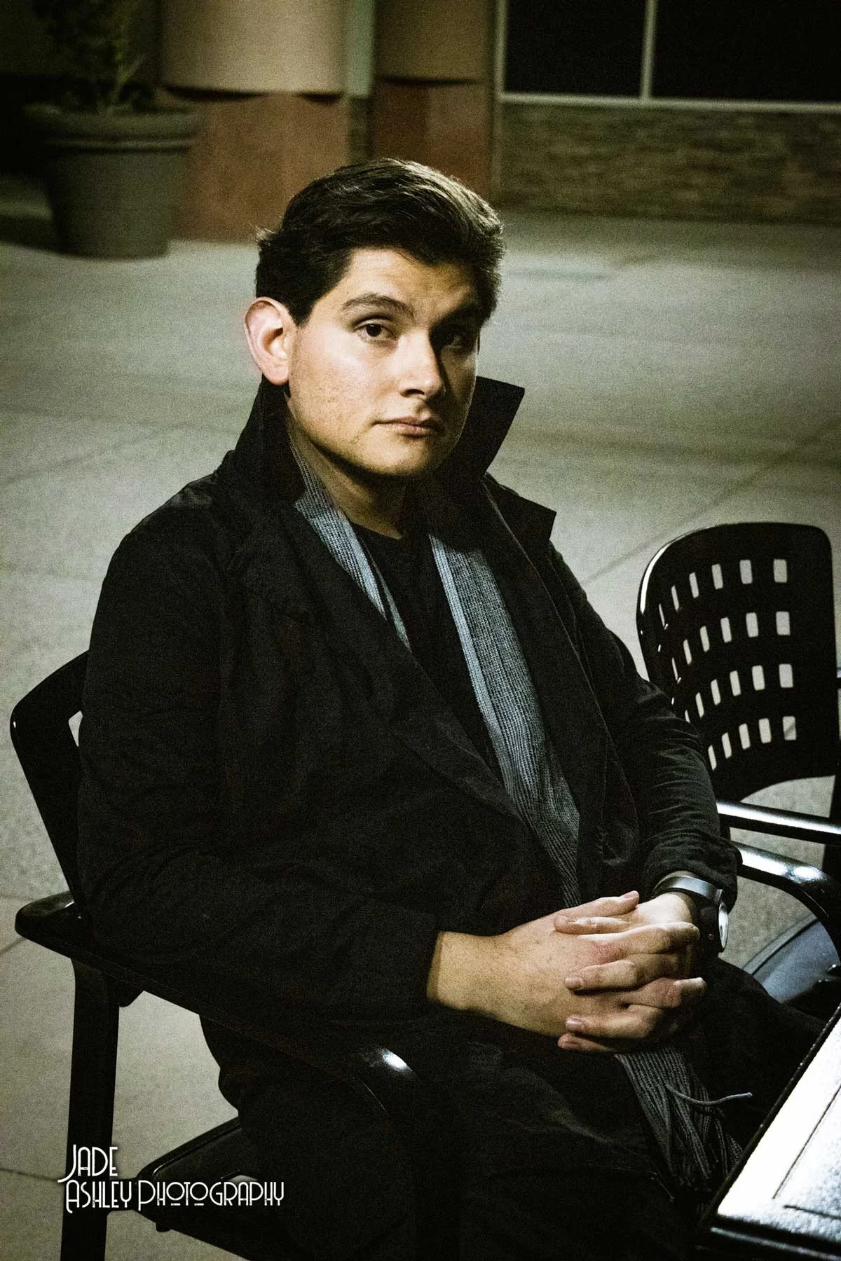 A young man with dark hair and light skin sitting outdoors at night, wearing a black jacket and a watch, with a serious expression.