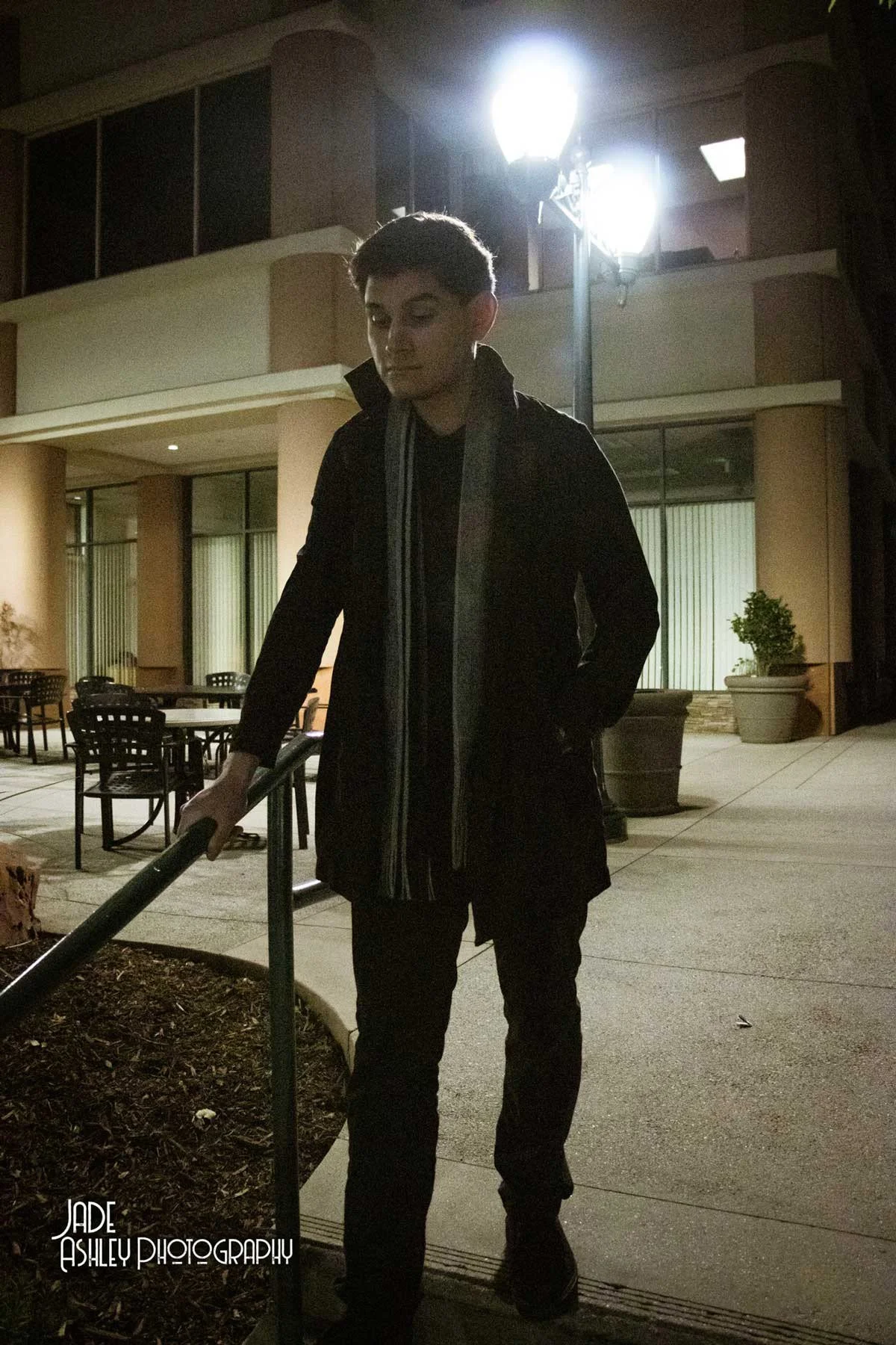 A young man wearing a dark jacket and striped shirt standing outside at night next to a metal railing, with a lit streetlamp and a building with large windows in the background.