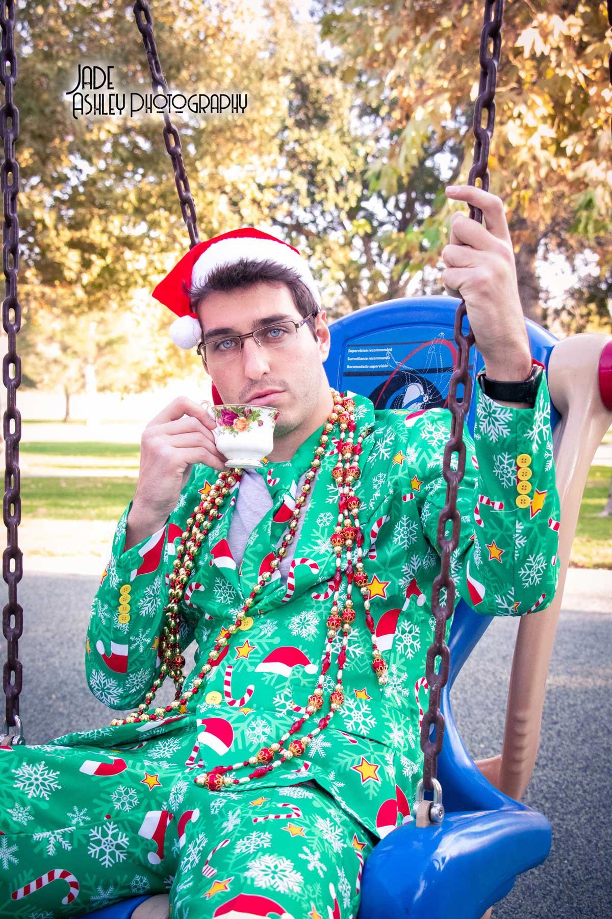 A young man wearing Christmas pajamas, a Santa hat, and multiple beads, sitting on a swing in a park. He is holding a teacup with a floral pattern and looking at the camera with a serious expression.