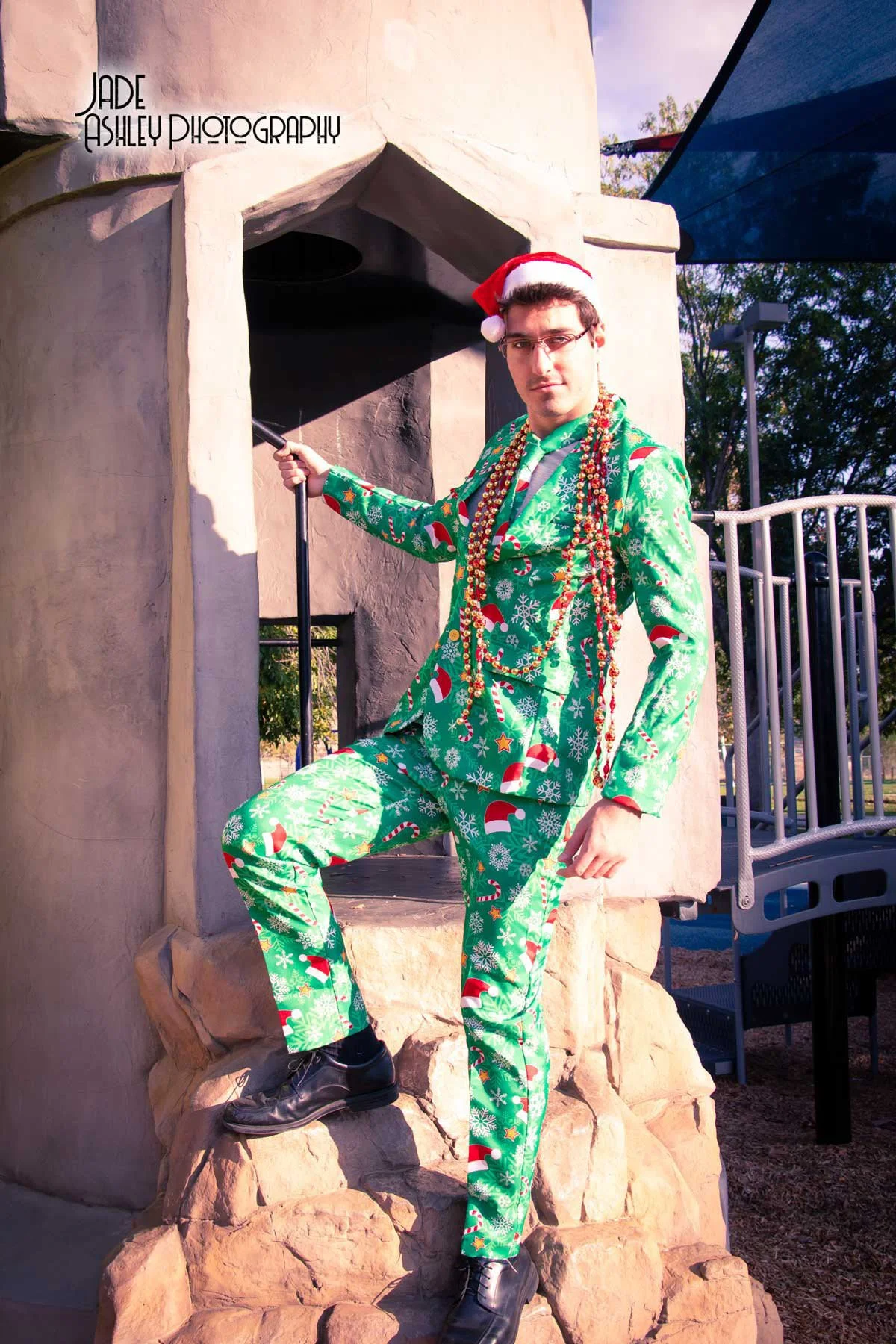 A man dressed in a Christmas-themed green suit with snowflakes, candy canes, and Santa hats print, wearing a Santa hat, standing on rocks beside a small structure, holding a stick, and adorned with Christmas beads. The sun is shining, with trees and 