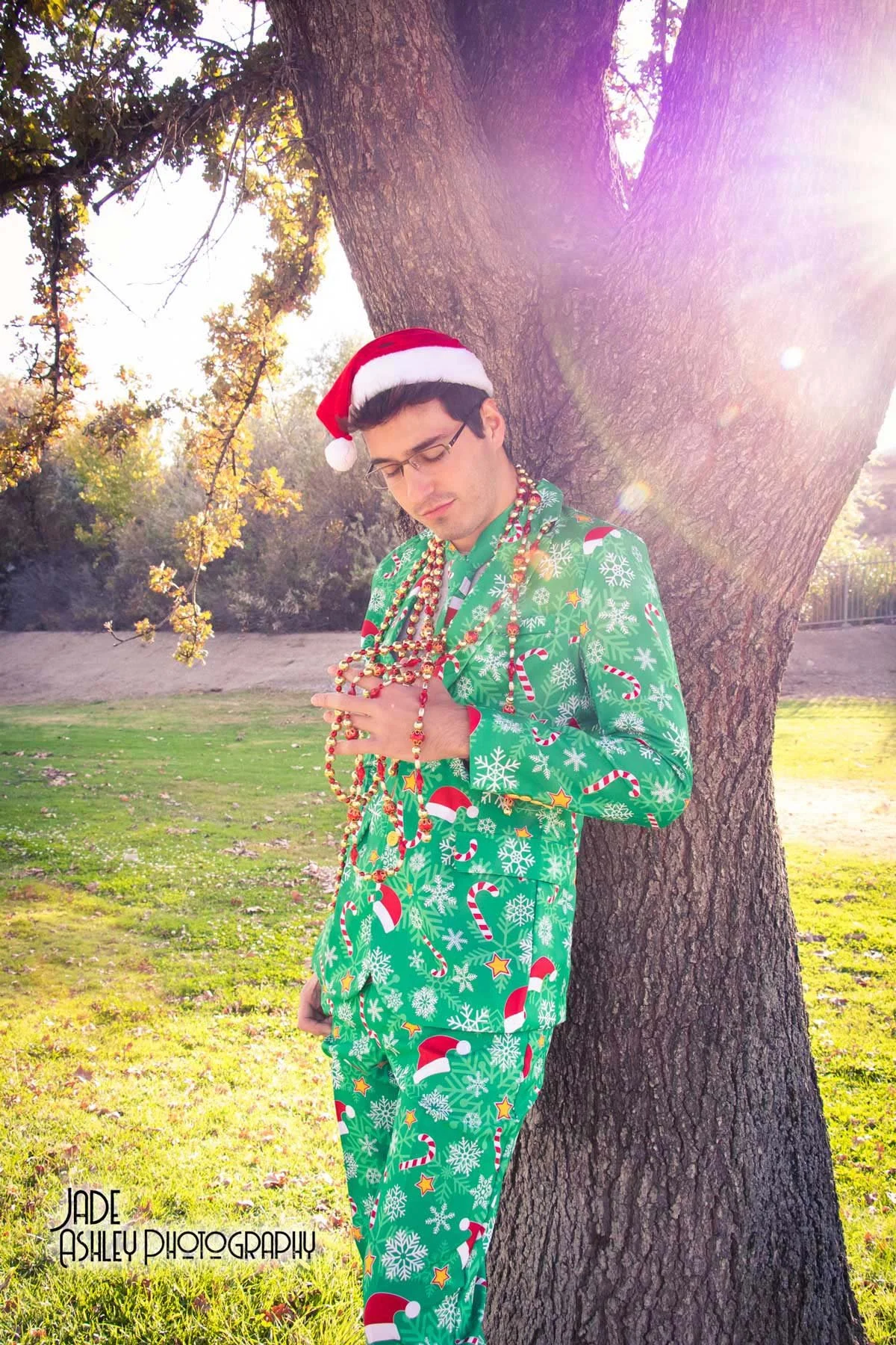 A man wearing festive green Christmas pajamas and a Santa hat is standing outdoors under a large tree with sunlight shining through the branches. He is holding and looking at a large amount of colorful beaded necklaces. The photo has a watermark that