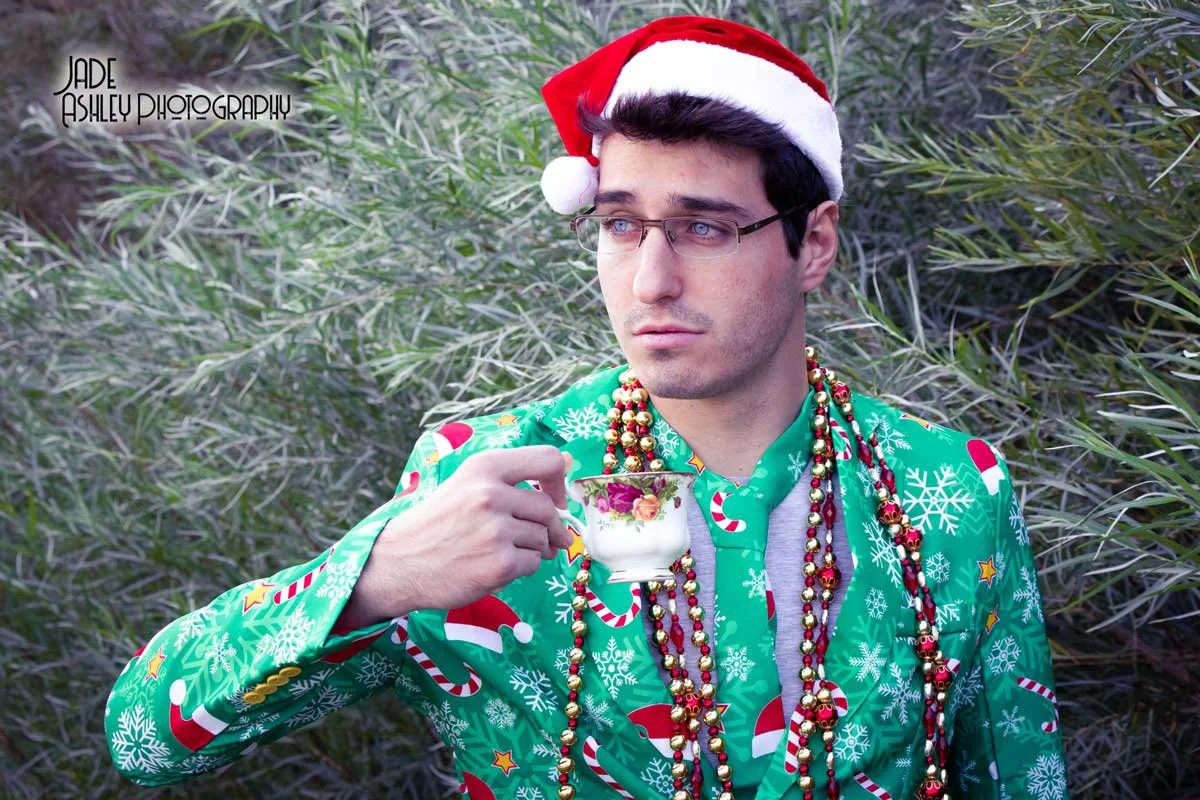 A young man wearing a Santa hat, glasses, and a festive Christmas-themed suit with candy canes and snowflakes, holding a teacup, and surrounded by holiday beads, standing outdoors among greenery.
