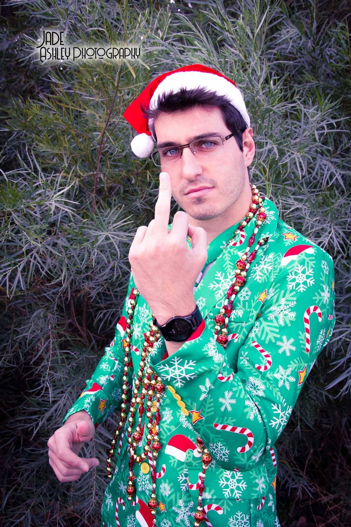 A young man with glasses, wearing a Santa hat and a festive green Christmas-themed shirt decorated with snowflakes, candy canes, and holly. He has multiple colorful bead necklaces around his neck, a black watch on his wrist, and is pointing his finge