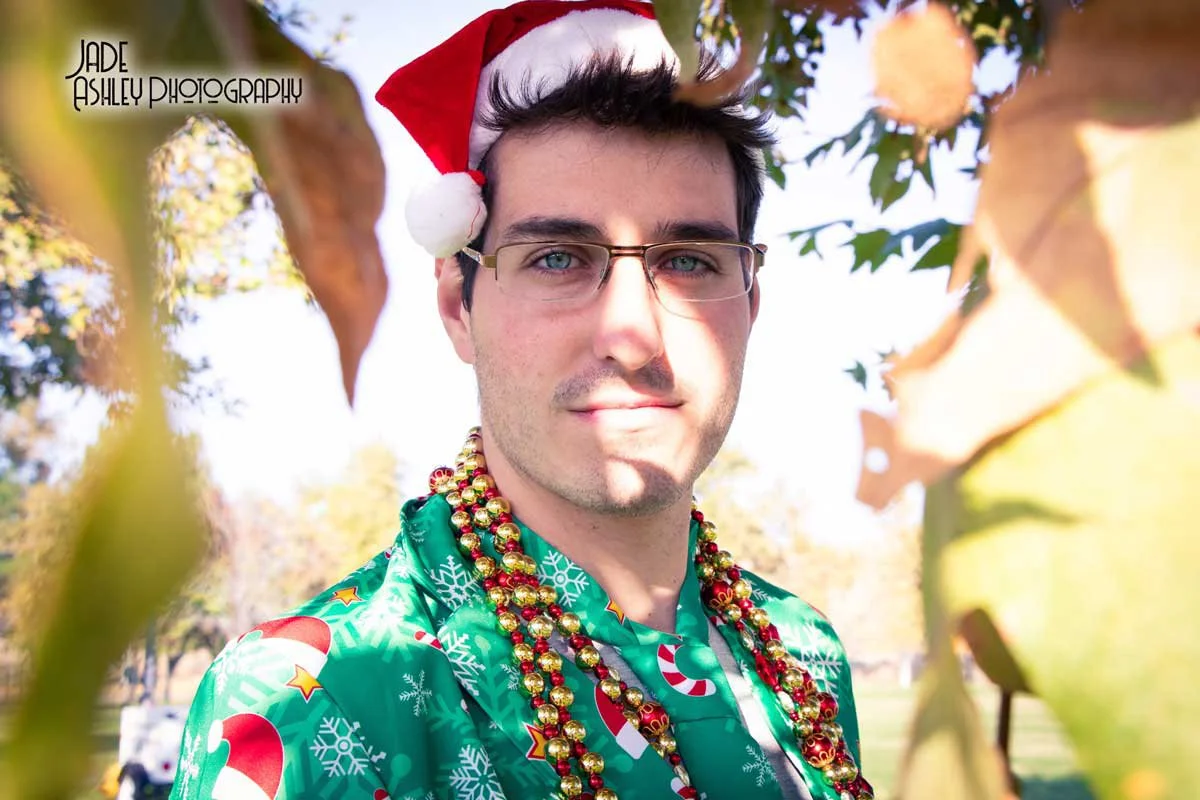 A man wearing glasses, a Santa hat, a festive green shirt with Christmas patterns, and bead necklaces, outdoors among autumn leaves with sunlight in the background.