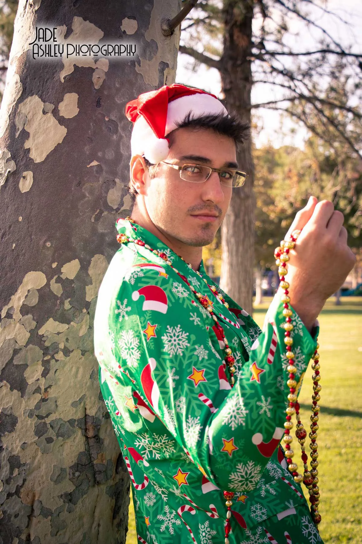 A man wearing a Christmas-themed jacket, Santa hat, and beaded necklace, standing outdoors against a tree in a park during daytime.