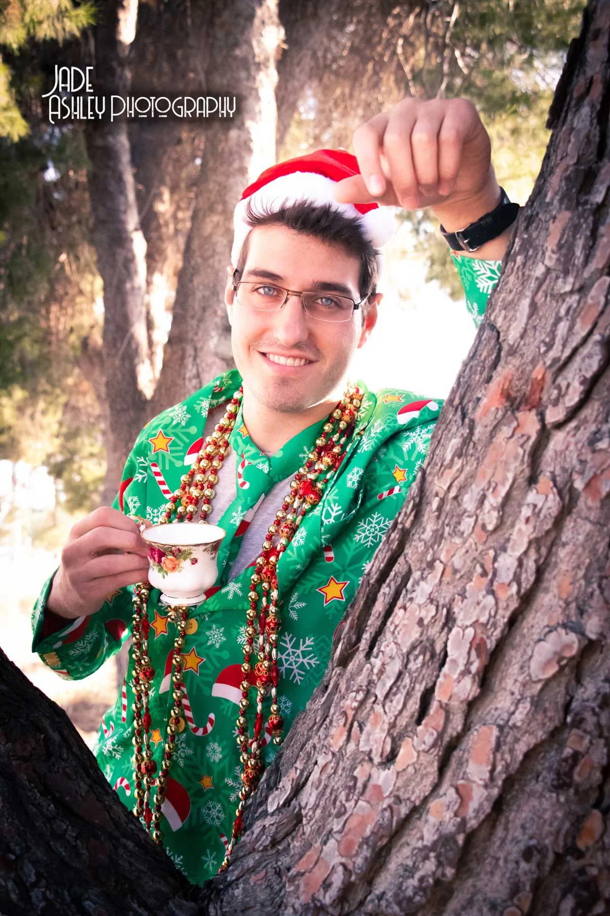 A young man wearing a Santa hat, glasses, and a festive Christmas sweater with candy canes, snowflakes, and stars, holds a floral teacup in one hand and smiles at the camera while leaning against a tree with a background of trees and sunlight.
