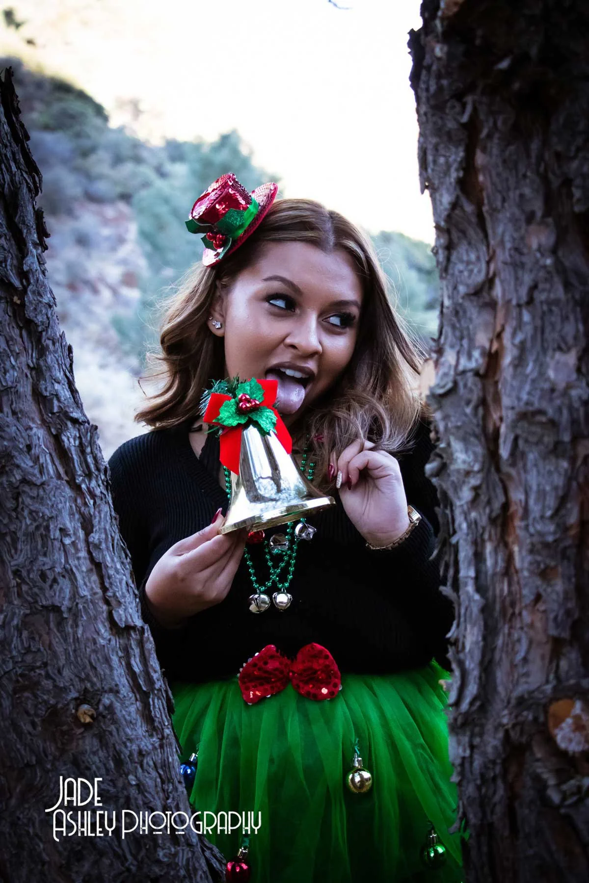 A woman with brown hair styled in loose waves is making a playful face, sticking out her tongue, and holding a Christmas bell ornament decorated with holly and red ribbons. She is wearing a red and green Christmas hat, green beaded necklace, and a gr