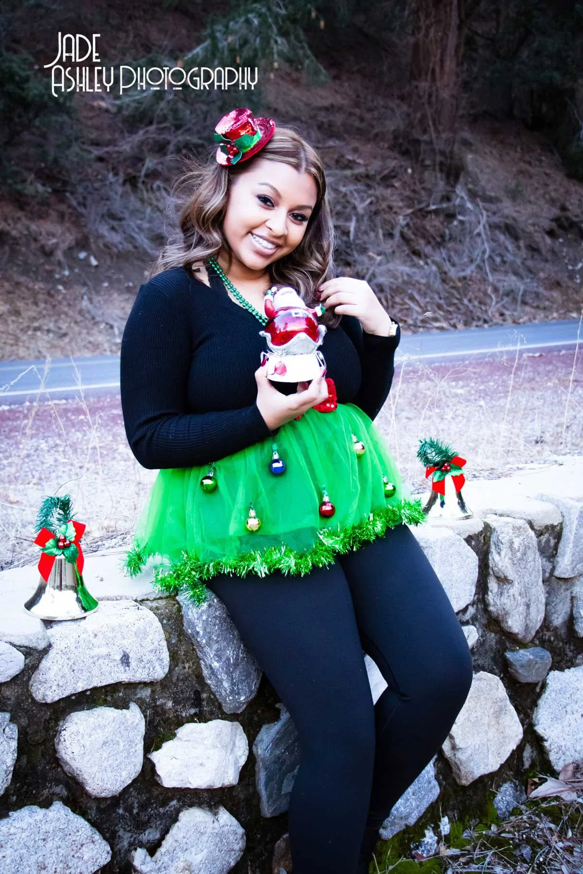 A young woman dressed in a festive Christmas outfit with a black long-sleeve top and a bright green tulle skirt decorated with Christmas ornaments, standing outdoors on a stone wall with Christmas bells and holly decorations nearby. She is smiling an
