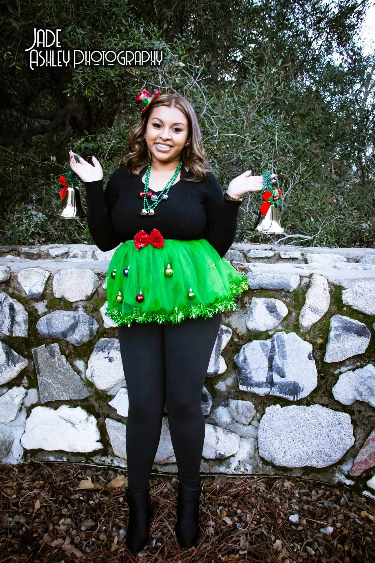Woman standing outdoors in front of a stone wall, wearing a Christmas-themed outfit with a green tutu, black top, black leggings, and black boots, holding small bells decorated with holiday ribbons in each hand. She has a small red bow on her tutu an