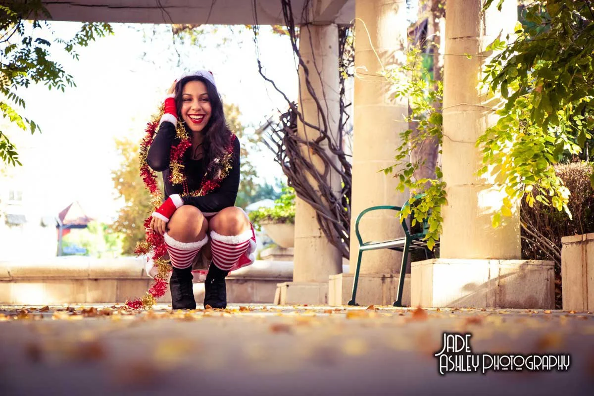 A young woman dressed in Christmas-themed clothing, including striped thigh-high socks, a garland around her shoulders, and a Santa hat, kneeling on the ground outdoors during autumn, smiling and talking on the phone.