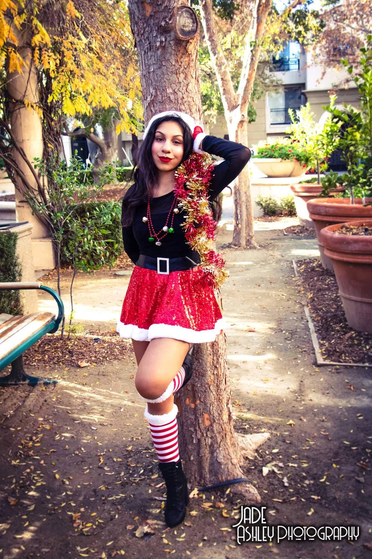 A woman dressed in festive holiday attire with a black long-sleeve top, red sequin skirt with white fur trim, striped knee-high socks, black boots, and holiday jewelry, standing outdoors against a tree with autumn leaves, smiling, with potted plants 