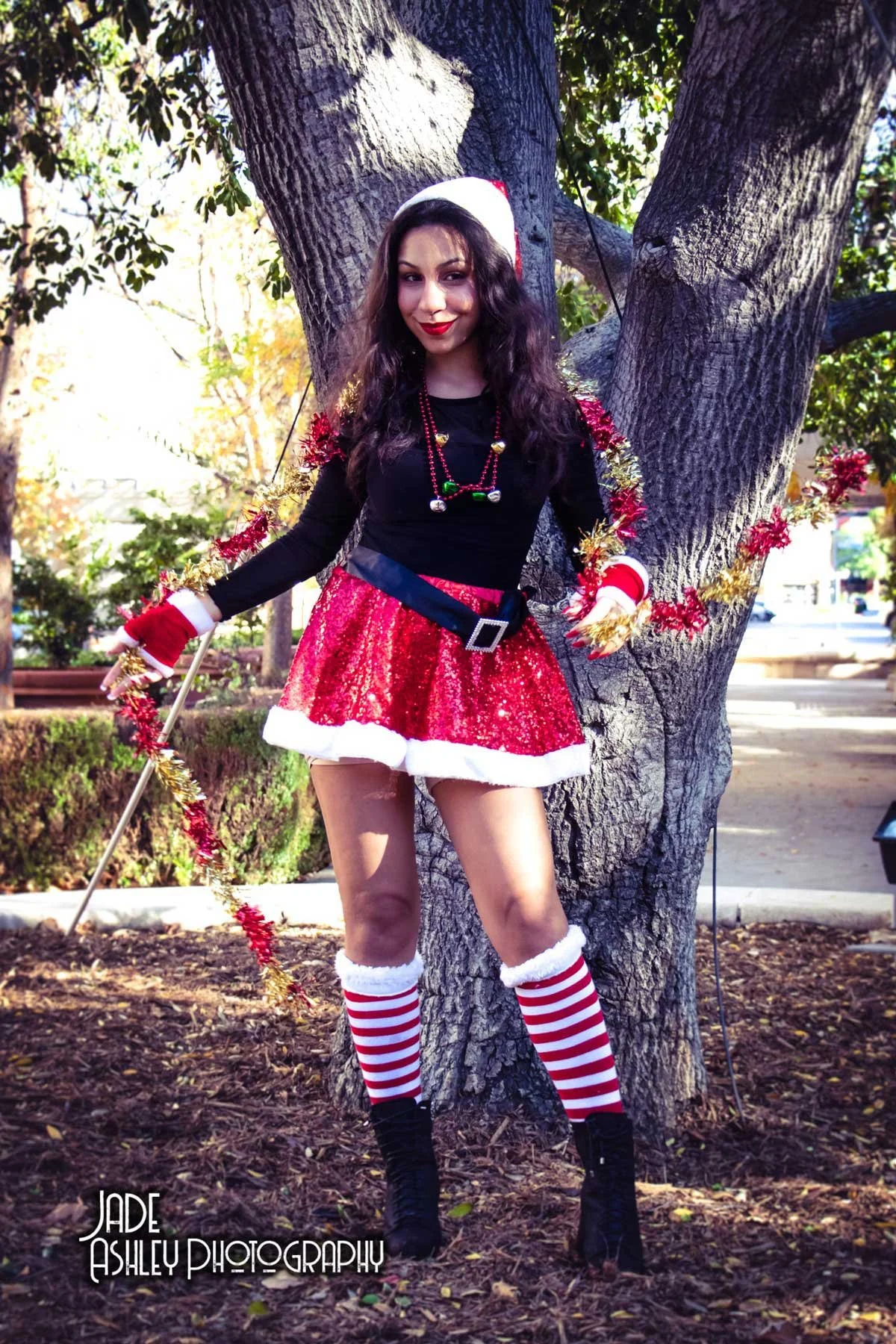 A woman dressed in a Christmas-themed outfit standing outdoors in front of a tree. She is wearing a black long-sleeve top, a red sequin skirt with white trim, striped red and white knee-high socks, black boots, and Christmas accessories including a S