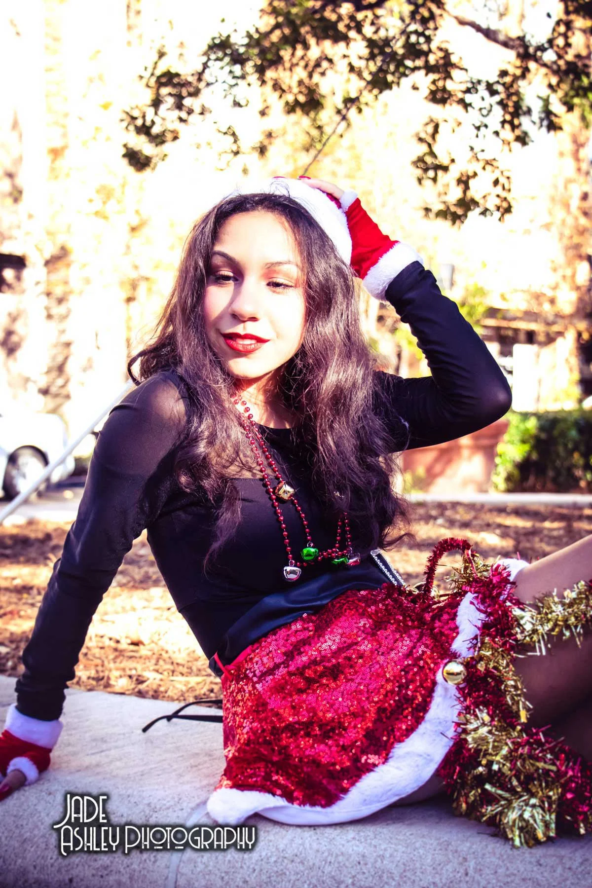 A woman sitting on the ground outdoors, wearing a red sequined skirt, black top, Christmas hat, and holiday accessories, with trees and sunlight in the background.