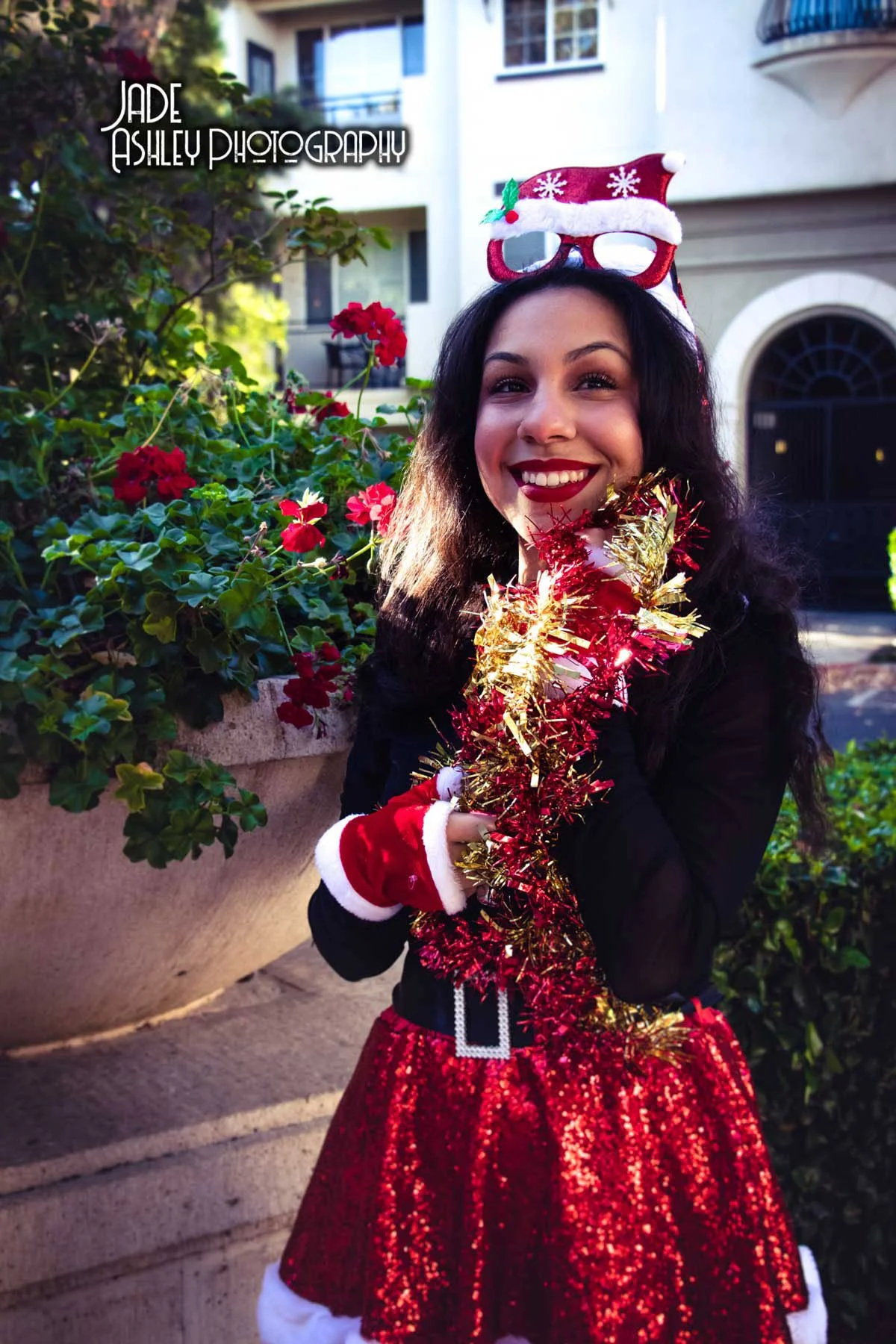 A young woman dressed in Christmas attire, wearing a red and glittery skirt, Santa hat glasses, red gloves, and holding gold tinsel, standing outdoors near a plant with red flowers, smiling at the camera.
