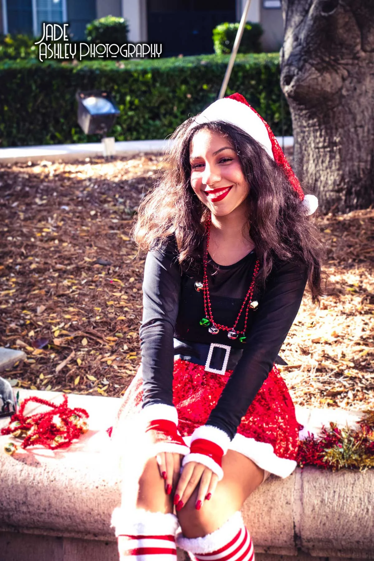 Young woman sitting outdoors on a ledge, dressed in Christmas-themed attire with a Santa hat, red velvet skirt, and striped knee-high socks, smiling at the camera.