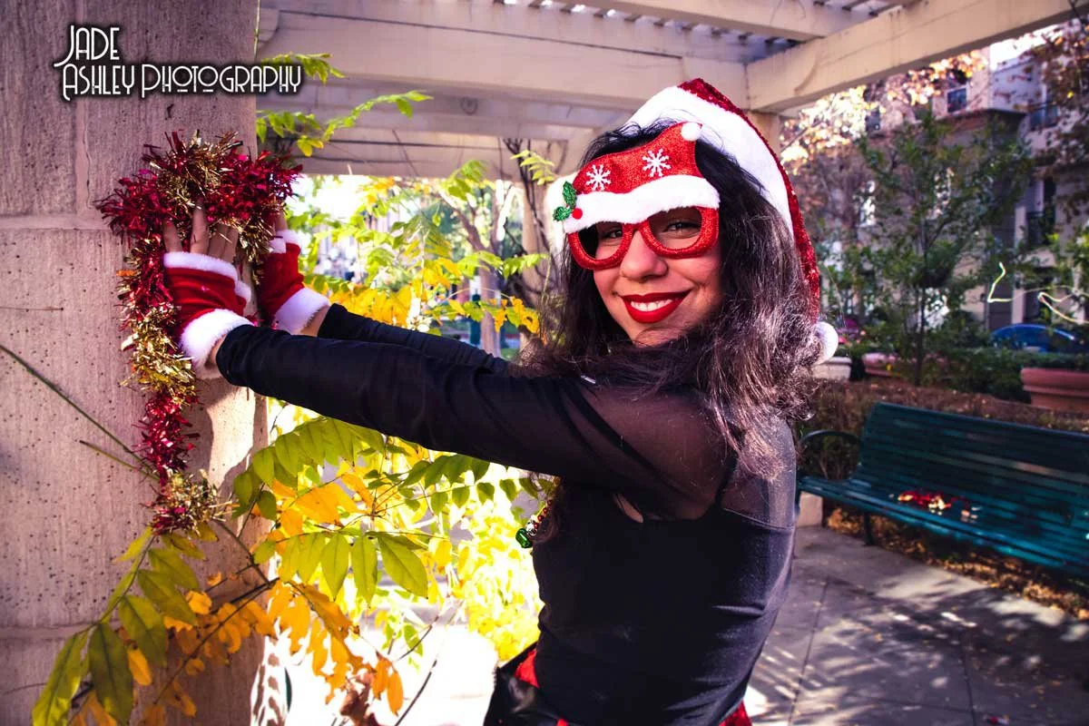 A woman wearing Christmas-themed accessories, including a Santa hat and red glasses with holiday decorations, is decorating a lamppost with red and gold tinsel during daytime in an outdoor park.