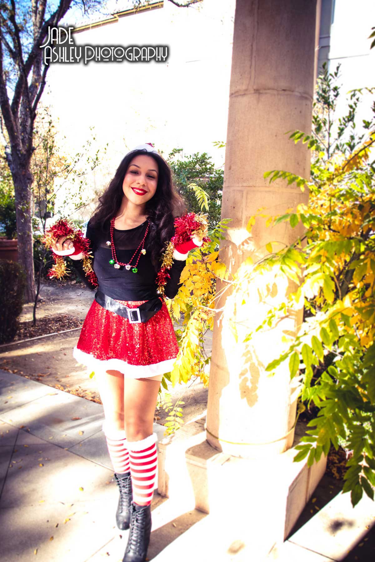 Woman dressed in Christmas-themed outfit, including a red sequined skirt with white trim, black top, striped red and white stockings, and black boots, standing outdoors next to a stone column and yellow-green foliage.