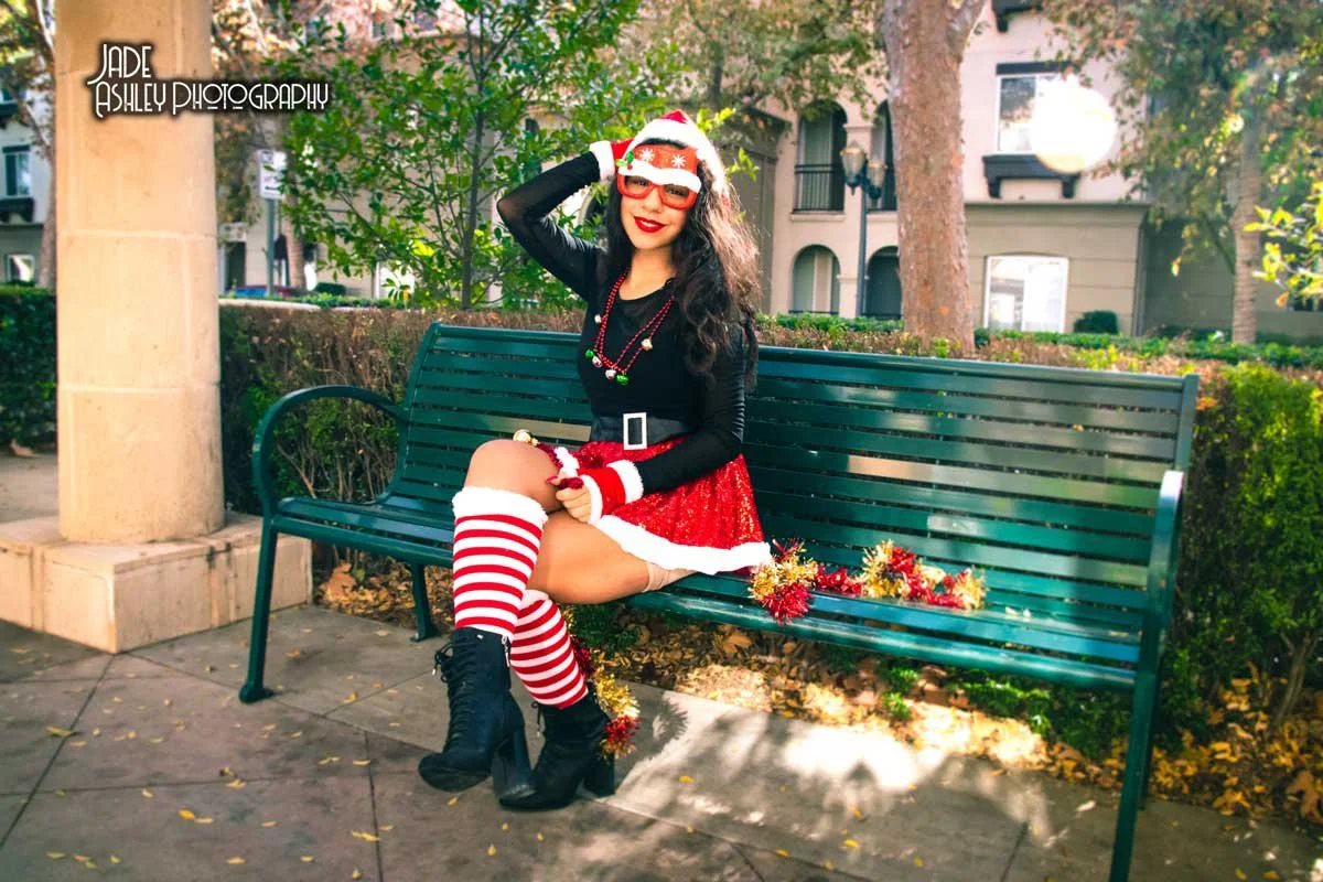 A woman dressed in a festive Christmas outfit sitting on a park bench, smiling, wearing a Santa hat, striped Christmas socks, black boots, and holiday jewelry, with Christmas decorations on the bench.