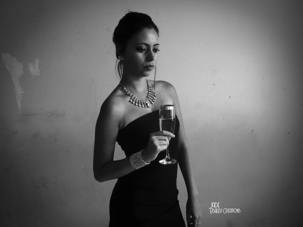 A woman in a strapless black dress holding a glass of champagne, wearing jewelry, looking down, with a plain wall background.
