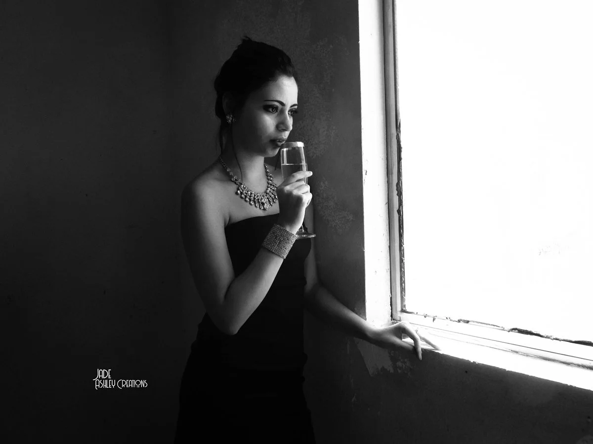 A woman in an elegant black dress and jewelry is looking out of a bright window while holding a glass of champagne.