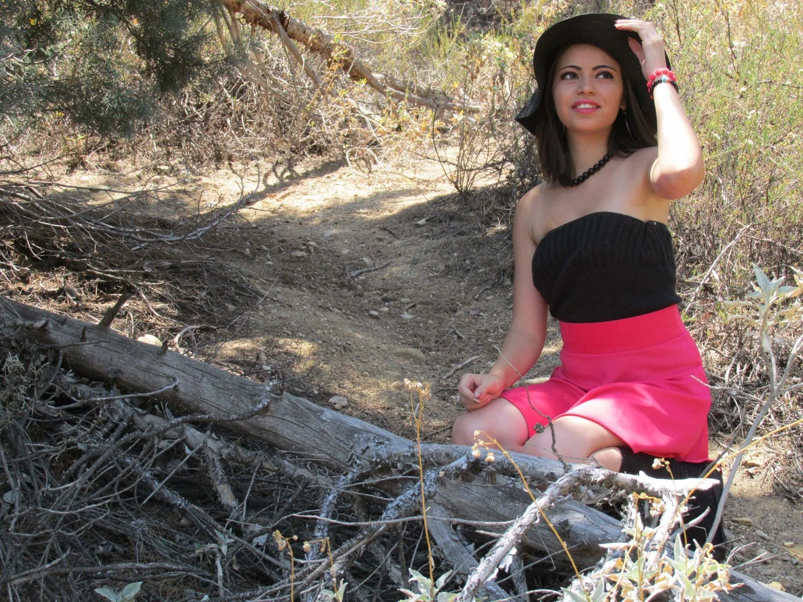 A woman kneeling on the ground in a dry, wooded area, wearing a black strapless top, pink skirt, a black wide-brimmed hat, and jewelry, looking into the distance with her hand on her hat.