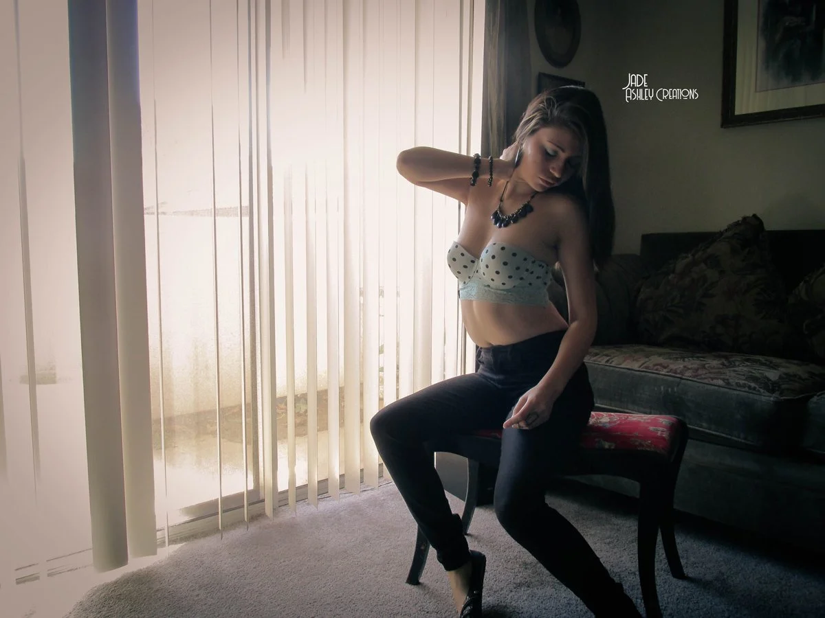 Woman with long hair posing indoors, wearing a polka dot strapless top, black pants, and jewelry, sitting on a chair near a window with vertical blinds.