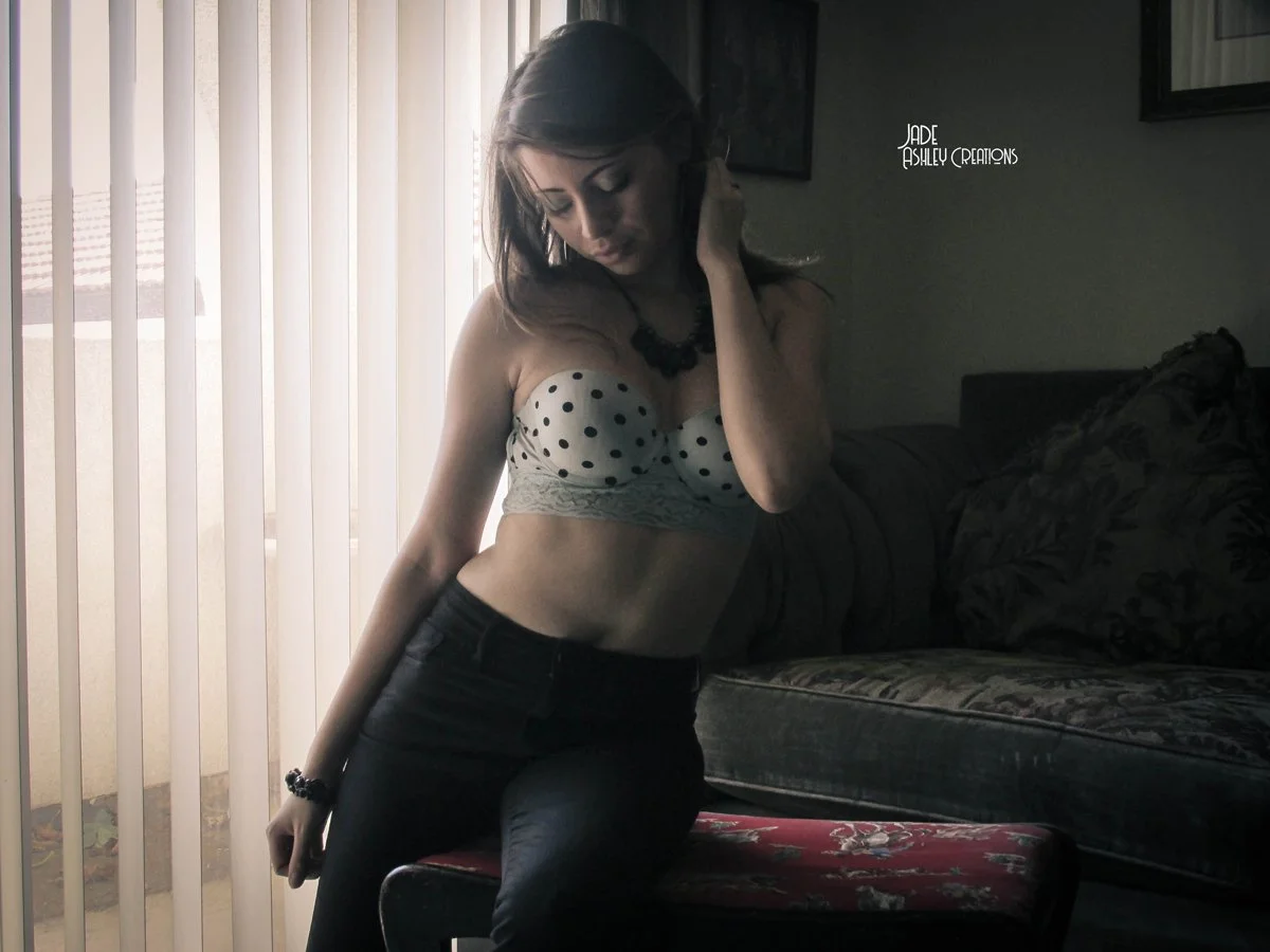 Young woman in polka dot top and black pants, posing indoors near a window with vertical blinds, with a sofa and picture frames in the background.