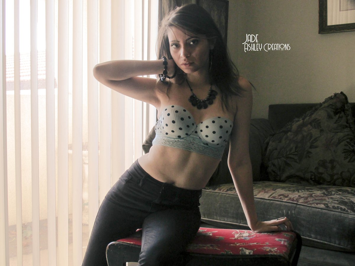 A woman with dark hair, wearing a polka-dot top and black pants, poses in front of window blinds and a couch, with jewelry including a necklace and bracelets.