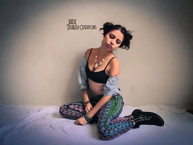 Young woman with dark hair styled in two buns, sitting on a white surface against a plain wall, wearing colorful patterned pants, a black crop top, gray jacket off her shoulders, and black boots, posing with a relaxed expression.