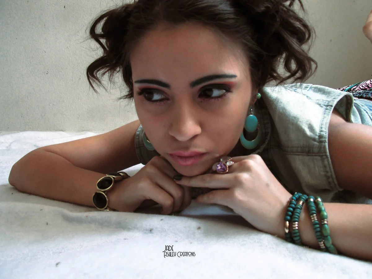 A woman lying on a bed, resting her chin on her hands, with short curly hair, wearing large turquoise earrings, a sleeveless denim vest, and turquoise jewelry, looking to her left.