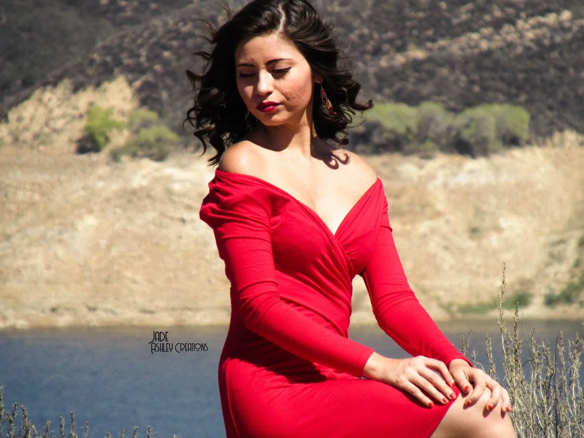 A young woman with dark, curly hair in a red off-shoulder dress sitting outdoors near a body of water, with a rocky hillside and sparse trees in the background.