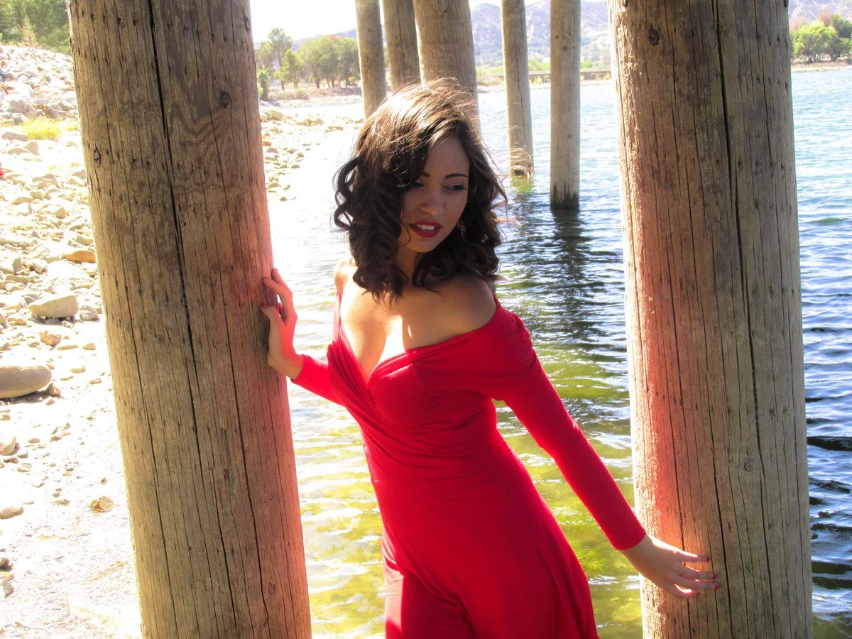A woman in a red dress leaning against wooden pilings at a lakeside under bright sunlight.