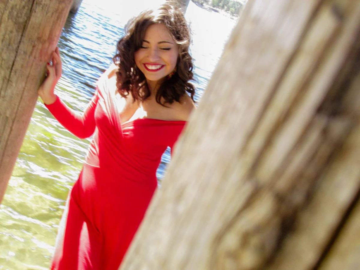 A woman with dark curly hair, wearing a red off-shoulder dress, smiling and posing between two wooden posts near a body of water.