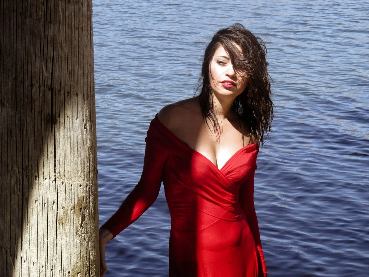 A woman with dark, wavy hair in a red dress standing by a wooden post near water.