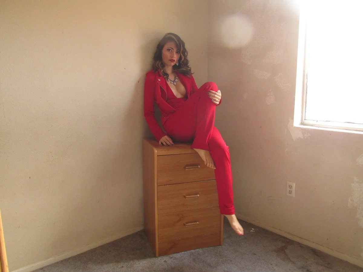 A woman dressed in a red outfit sitting on top of a wooden dresser in a sparsely decorated room with beige walls and a window.