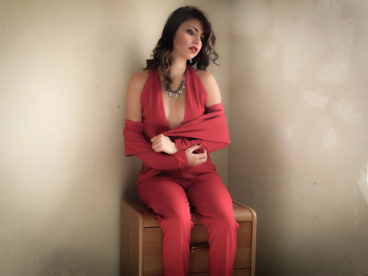 A woman with dark hair and makeup, wearing a red dress and necklace, sitting on a wooden box in a corner, looking to the side with a contemplative expression.