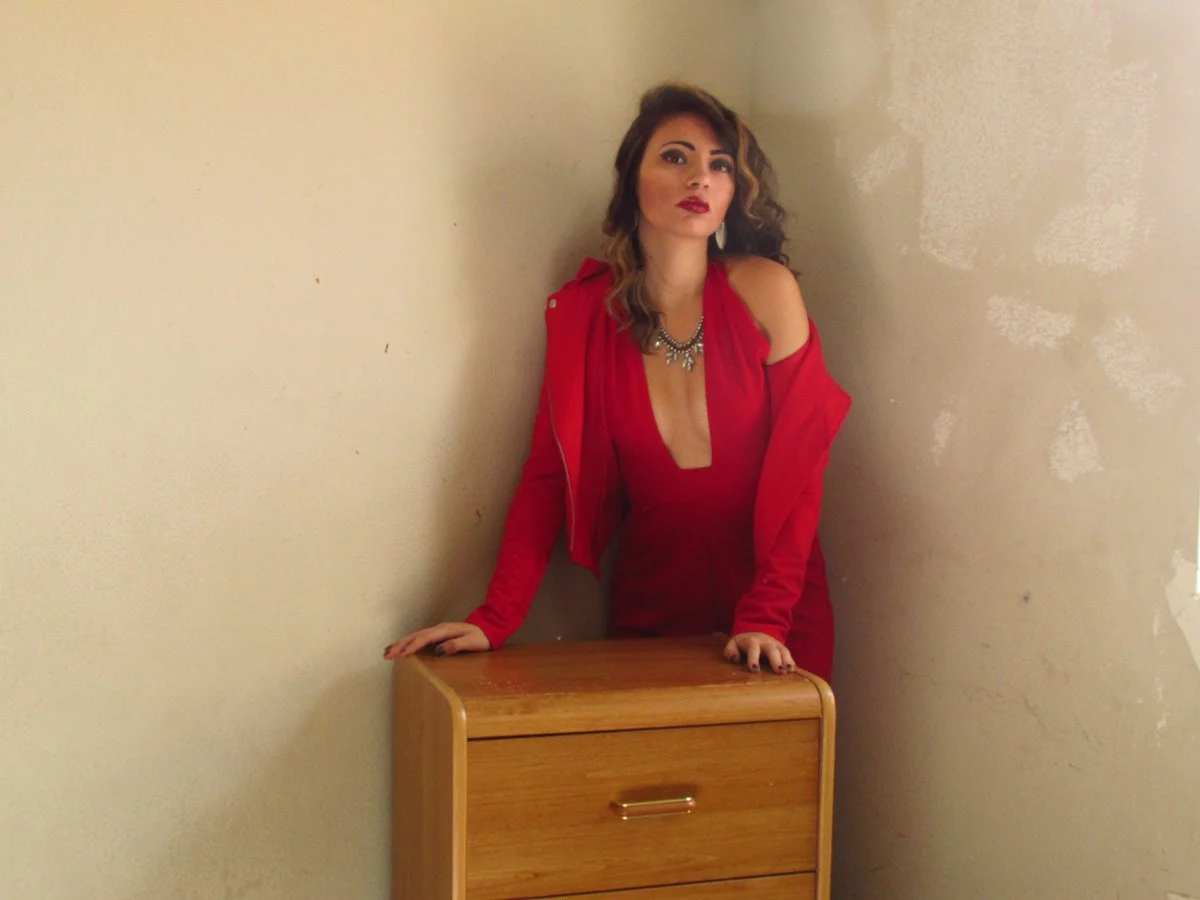 Woman in a red dress and jacket standing behind a small wooden dresser, leaning on it with one hand, posing against a beige wall.