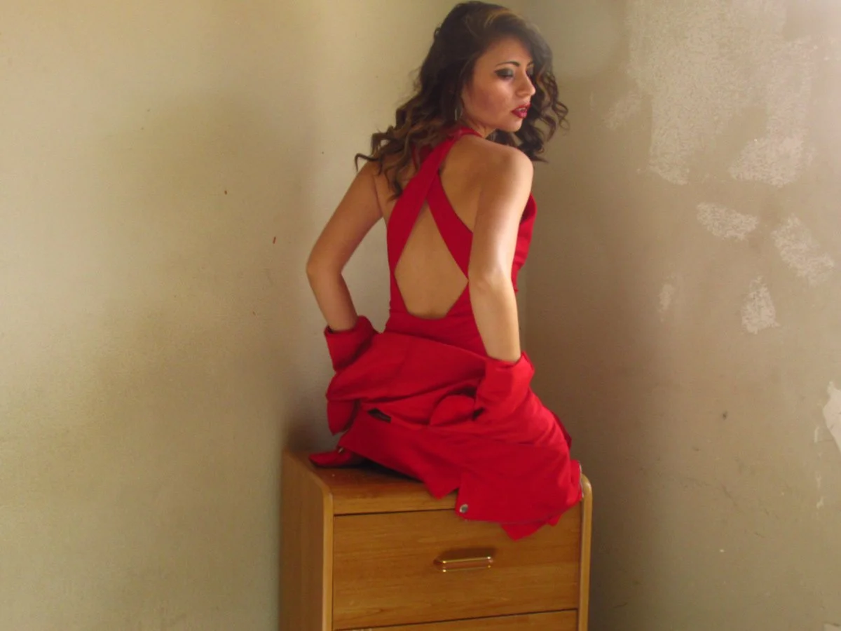 Woman in a red dress with an open back, sitting on a wooden dresser against a beige wall.